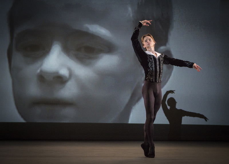 Daniil Simkin is in relve with his right arm above his head while a projection of a photo of him as a child is displayed behind him